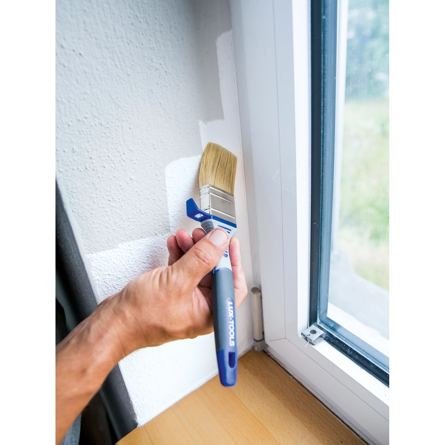 Hand streicht mit LUX Flachpinsel (30mm) eine Wand. Der Pinsel hat einen Eimerhaken.