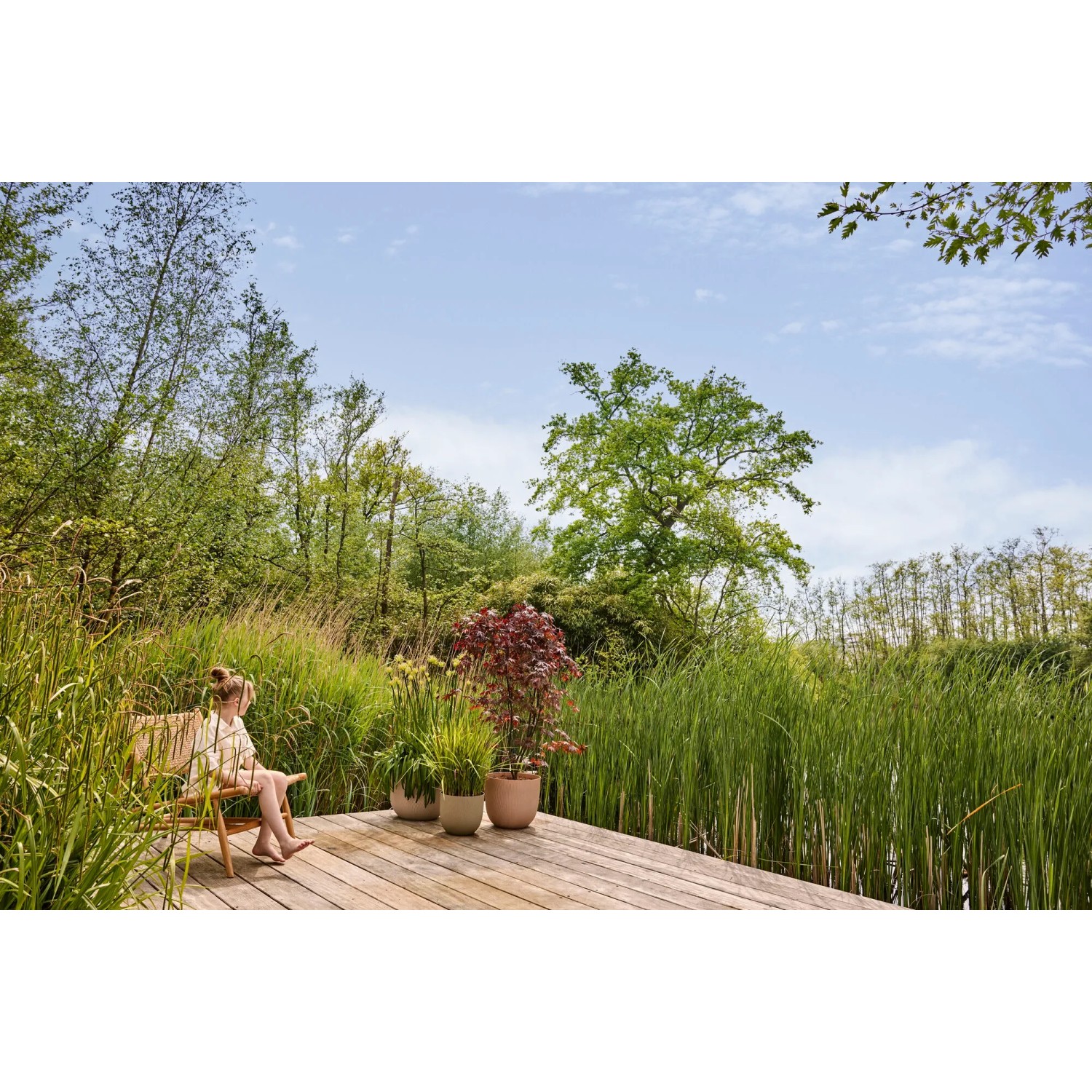 Elho Blumentopf Sereh, Ø 47 cm, grün, auf Holzterrasse mit Bepflanzung und sitzender Person.