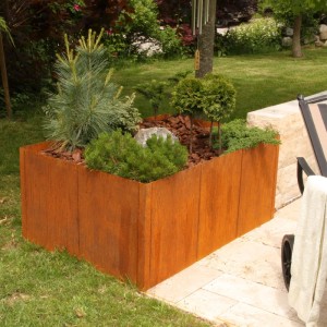 Rechteckiges Corten-Stahl Hochbeet im Garten, bepflanzt mit Nadelgehölzen und Rindenmulch. Modularer Bausatz.
