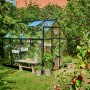 Schwarzes Halls Gewächshaus Qube 68 (5 m²) im Garten mit Pflanzen und Sitzgelegenheit.