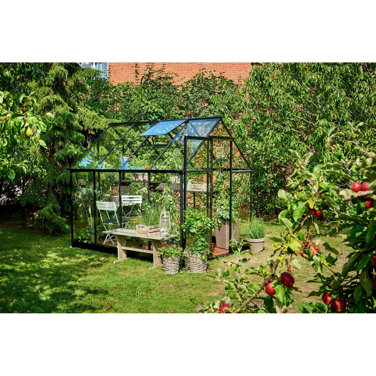 Schwarzes Halls Gewächshaus Qube 68 (5 m²) im Garten mit Pflanzen und Sitzgelegenheit.