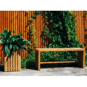 Lackierter Sichtschutzzaun Quadra (180x180cm) in Natur mit Bank und Pflanzenkübel vor grünem Bewuchs.