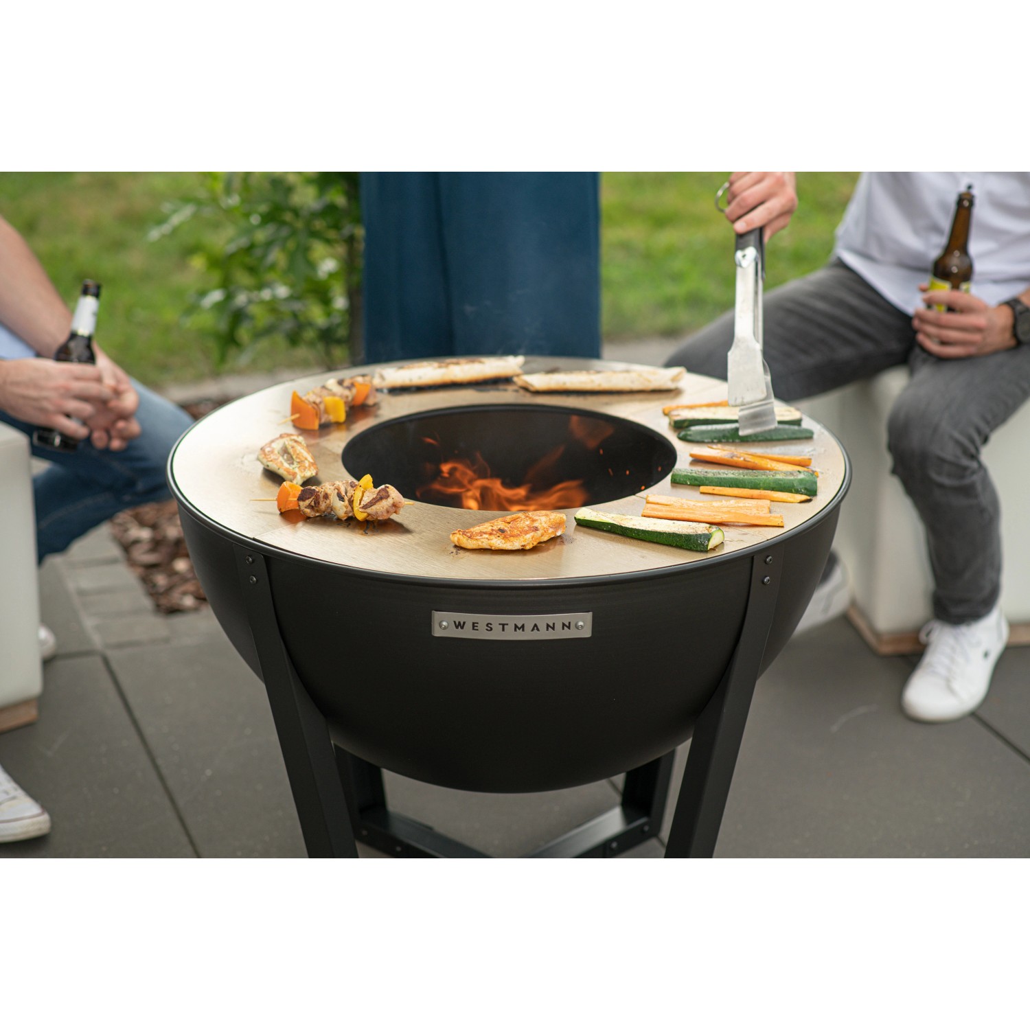 Westmann Premium Grill Feuerstelle, Ø 81cm, schwarz, mit Grillgut und Personen im Garten.