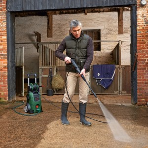 Mann reinigt mit Bosch Hochdruckreiniger AdvancedAquatak 140 eine Fläche vor einem Stall.