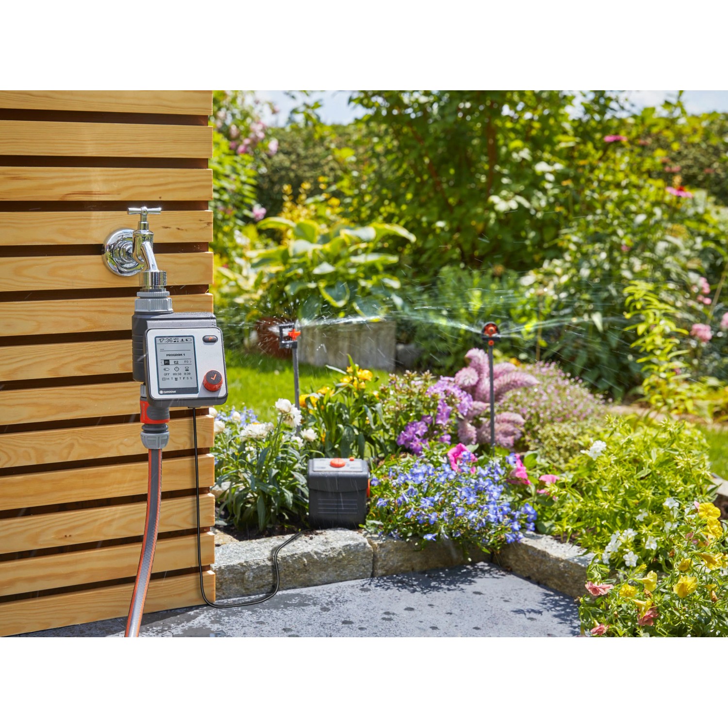 Gardena Bewässerungscomputer Master am Wasserhahn mit Gartenbewässerung im Hintergrund.