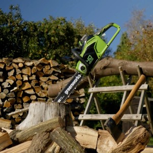 Greenworks Akku-Kettensäge GD40CS18 (40V Solo) beim Holzsägen mit Axt und Holzstapel im Hintergrund.