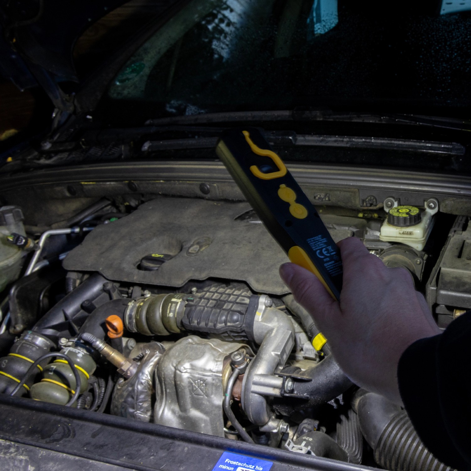 LED's Work Akku Handlampe leuchtet in einem Motorraum. Der LED-Baustrahler ist schwarz-gelb.