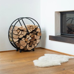 Schwarzes, rundes Westmann Kaminholzregal Osiris aus Stahl, gefüllt mit Holzscheiten, neben Kamin.