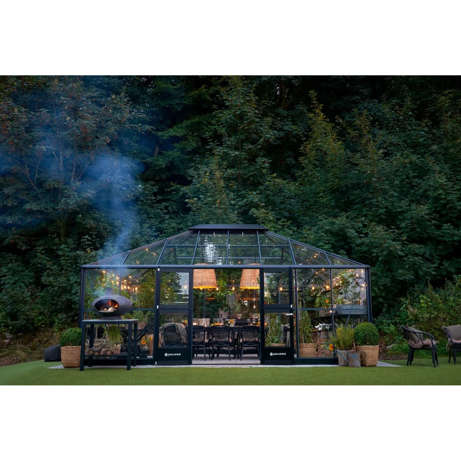 Juliana Gewächshaus Grand Oase 25 in Schwarz-Anthrazit mit Esstisch und Pizzaofen im Garten.
