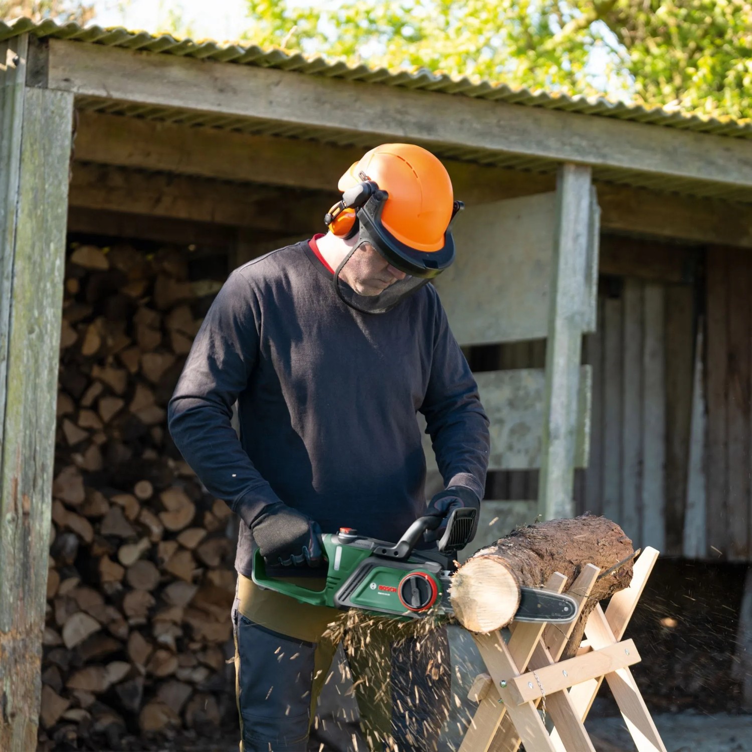 Mann sägt Holz mit Bosch Akku-Kettensäge PKE18V-30-28. Inkl. Helm und Sägespäne.