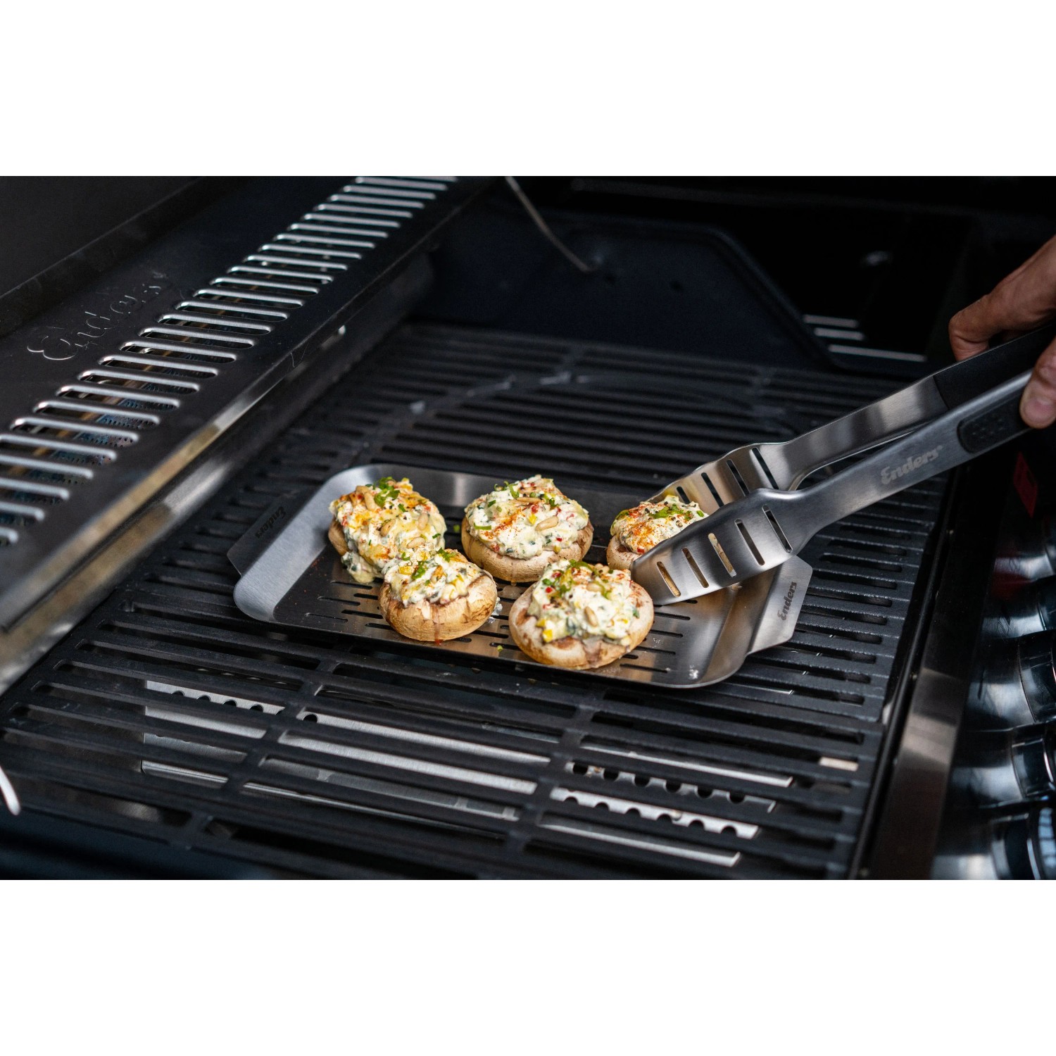 Enders Grillschale aus Edelstahl mit gefüllten Champignons und Grillzange auf einem Grill.