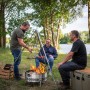 Petromax Dreibein mit Grillrost über Feuerstelle, umgeben von drei Personen in der Natur.